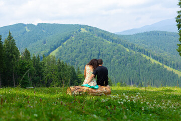 Young couple in love hugs sitting on the log and enjoy betiful of nature mountain lanscape overgrov with green trees