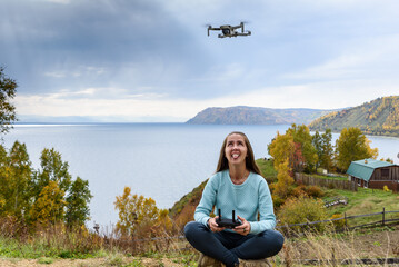 Wall Mural - Beautiful young woman having fun with a mini drone outdoors in Lotus pose. The girl sits against the autumn forest background