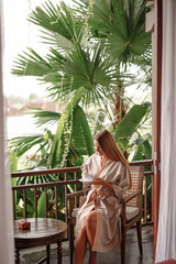 Wall Mural - Girl in bathrobe with cup of coffe at the jungle balcony sit on the chair, enjoy morning in tropics