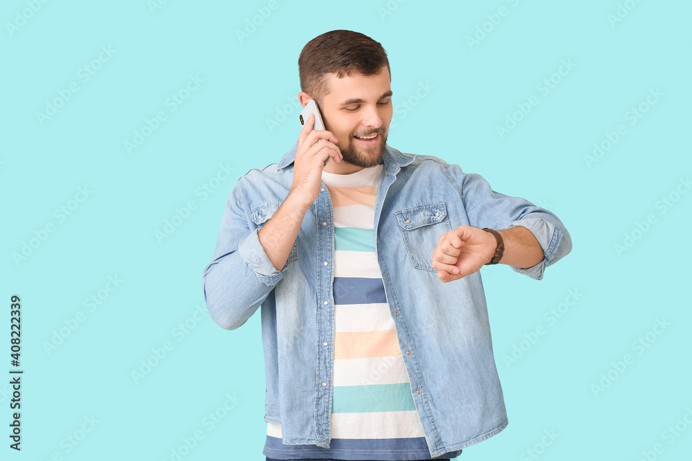 Handsome man looking at wristwatch and talking by mobile phone on color background