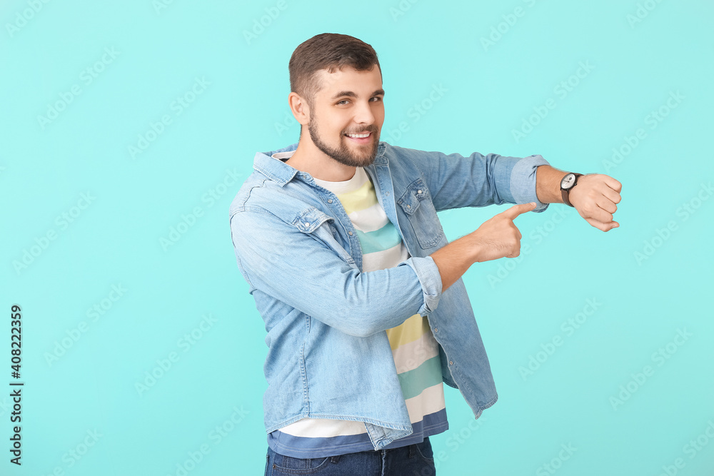 Handsome man pointing at wristwatch on color background