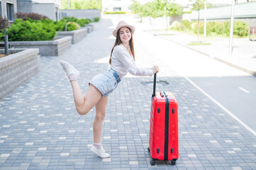 Young caucasian woman in a hat and shorts stands on the street with a red suitcase.