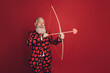Photo of positive retired cherub man hold bow arrow prepare shoot wear heart print tux isolated red background