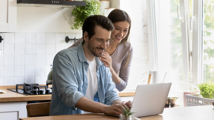 Sharing good news. Happy smiling millennial man use laptop at home office on kitchen typing pleasant message. Caring young woman wife standing by hugging husband with warmth reading email from screen