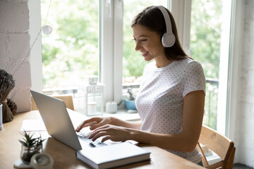 Wall Mural - Happy millennial female sitting at desk in home office studying working online using laptop earphones. Positive young lady wearing headphones communicating in virtual web conference via social media