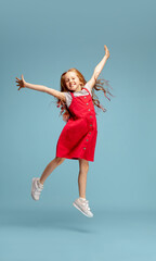 Wall Mural - Weightless, freedom. Happy redhair girl in red dress on blue studio background. Looks happy, cheerful. Copyspace for ad. Childhood, education, emotions, facial expression concept. Jumping high, flying