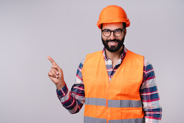 Wall Mural - Cheerful Arab builder in hardhat and uniform pointing at copy space isolated over grey background