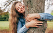 © iuricazac - Candid portrait of a joyful young woman smiling, wearing a blue denim shirt embraces a tree, posing on nature background. Gorgeous female enjoying the time in the park. Mental health concept