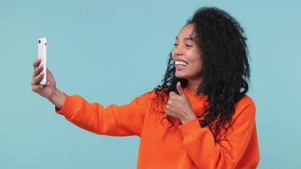 Wall Mural - Young african woman with in orange sweatshirt get video call using mobile cell phone doing selfie videoconference talking conducting pleasant conversation greets with hand isolated on blue background