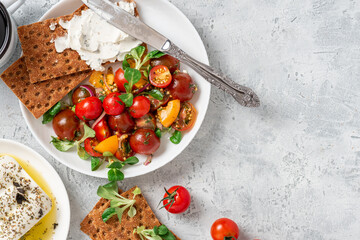 Sticker - Tasty breakfast or lunch. Salad of colorful cherry tomatoes, onions, herbs, and olive oil, served with crispbread and cream cheese on a gray background top view. Free space for text.