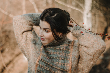 Wall Mural - Young woman wearing warm cosy jumper, tying hair into a bun, watching to the side smiling, in an autumn forest background