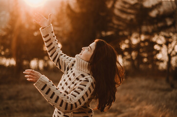 Wall Mural - Warm portrait of a young woman wearing a comfy sweater waving arms at sunset light with trees and lake in the background