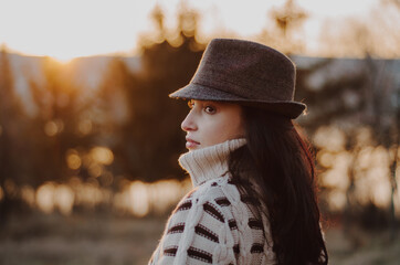 Wall Mural - Portrait of young woman in warm sweater wearing a hat looking to the side, in a forest at sunset