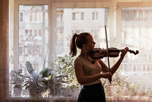 Silhouette Of A Young Girl, A Musician. Playing The Violin In The Background Of The Window