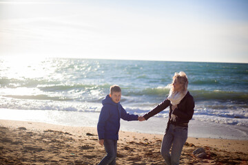 Happy family, mother with son walking wirh fun in the sea shore on windy day. People dressed warm clothes.