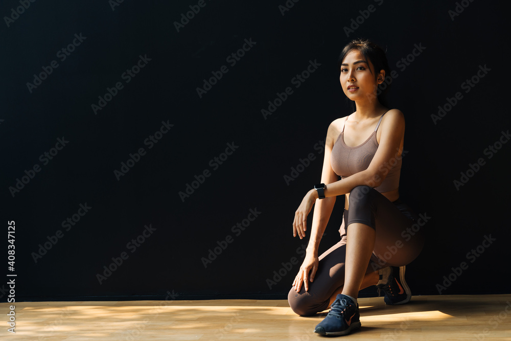 Focused young Asian sportswoman relaxing and getting ready for workout while crouching in studio on black background - full body length - obrazy, fototapety, plakaty 