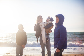 Happy family, mother with sons walking wirh fun in the sea shore on windy day. People dressed warm clothes.