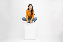 Smiling Young Asian Woman Sitting On White Box And Pointing Down Empty Copy Space Isolated On White Background