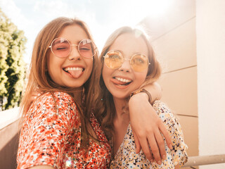 Two young beautiful smiling hipster female in trendy summer sundress.Sexy carefree women posing on the street background in sunglasses. They taking pov selfie  photos on smartphone at sunset