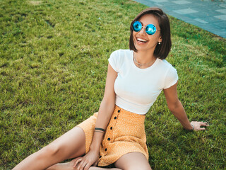 Wall Mural - Portrait of beautiful smiling brunette model dressed in summer hipster clothes. Trendy female sitting on the grass in the park. Funny and positive woman having fun