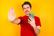 © Roquillo - Young Caucasian man wearing red t-shirt standing against yellow background using and texting with smartphone with open hand doing stop sign with serious and confident expression, defense gesture