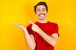 © Roquillo - Young Caucasian man wearing red t-shirt standing against yellow background pointing and holding hand showing adverts