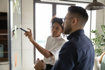 Close up diverse colleagues developing plan, confident African American businesswoman mentor coach writing idea or task on glass wall, employees working on project strategy in creative office room