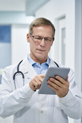 Mature doctor wearing glasses using digital tablet in hospital. Old senior male physician holding pad computer e health technology consulting patients online. Medical telehealth, tele medicine concept