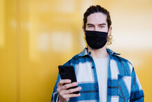 Young Man With Protective Face Mask And Mobile Phone Staring While Standing Against Yellow Wall