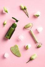 Jade Stone Face Roller, Gua Sha, Face Cream Lotion On Pink Background With Ice Cubes And Frozen Flowers. Facial Skin Treatment Concept