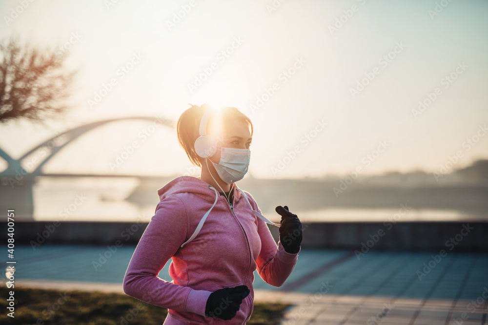 Beautiful young and fit woman in good shape is running and jogging alone on city bridge street. She is wearing protective face mask to protect herself from virus or allergy infection.  - obrazy, fototapety, plakaty 
