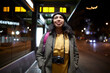 © Cavan Images - woman standing with a camera waiting at the bus station at night
