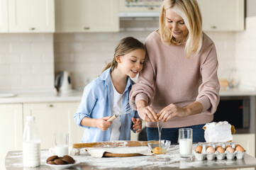 Caucasian happy mom and her beloved little daughter spend time together in the kitchen. Grandma teaches her cute granddaughter to cook pie, they are happy to cook together, having fun and laughing