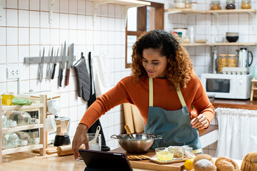 Happy young African American woman cooking in kitchen follwing online recipe in digital tablet, home hobby concepts