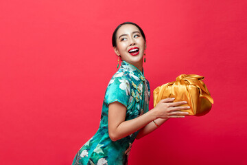 Smiling pretty Asian woman wearing traditional dress holding golden gift box looking at copy space aside in isolated red studio background for Chinese new year concepts