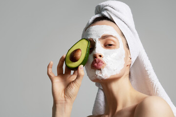 Funny young woman in mask and white towel holding piece of avocado isolated on gray background. 