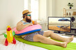 Funny young man with sunglasses and inflatable beach toys sipping cocktail and watching travel show on TV. Concept of canceled summer holiday plans, vacation in lockdown at home or Covid-19 quarantine