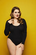 Plus-size model woman with bright makeup in black bodysuit over yellow background, isolated. Young sexy plump woman
