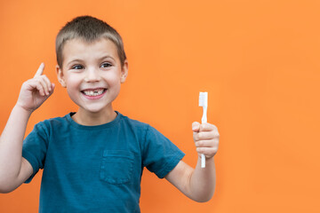 Boy without milk upper tooth in blue t-shirt holds toothbrush in hand on the orange background. Copy space.