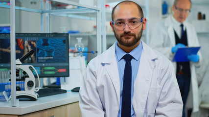 Tired doctor looking exhausted at camera in modern equipped lab. Multiethnic team examining virus evolution using high tech and chemistry tools for scientific research, vaccine development.