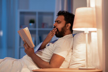 People, Bedtime And Rest Concept - Tired Indian Man Reading Book And Yawning In Bed At Home At Night