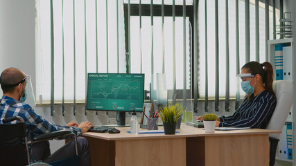 Paralysed financial worker comming in wheelchair in new normal business office with protection mask waving at colleague. Immobilized freelancer in corporate company respecting social distance.