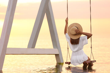 Wall Mural - Young woman enjoying sunrise on swing over water