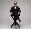 © opolja - Portrait of a respectable mature man in a suit and sunglasses sitting on chair on a gray background