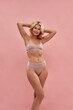 Feeling good. Happy charming middle aged caucasian blonde woman in underwear smiling at camera, keeping arms raised while standing against pink background
