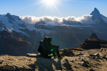 Young romantic Asian couple traveler sitting hugging on a cliff admiring the amazing landscape of Matterhorn mountain peak with snow-capped penetrated the white cloud in a clear blue sky with sunshine