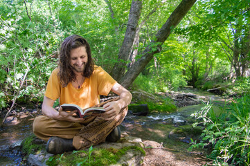 Young man with long hair sitting cross legged on a moss covered stone on the shore of a river in the woodlands reading a book and smiling.