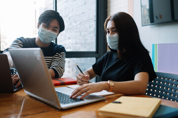 Asian woman and man workers meeting together with laptop for financial and wear protective masks prevent corona virus or covid19 at co working space .Health and teamwork concept.