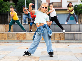 Cute blonde preteen b-girl dancing with group of friends on city street. Urban lifestyle. Hip-hop generation