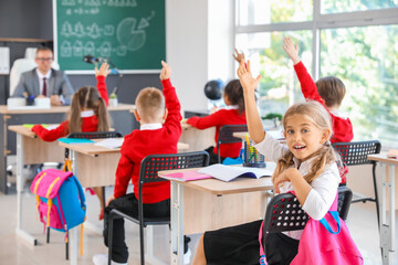 Cute schoolgirl with raised hand during lesson in classroom